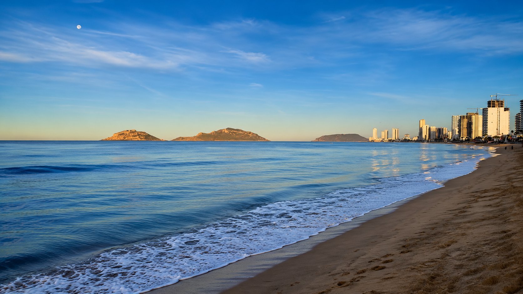 Scenic beach with white sand and breaking waves, high-rise buildings along the shoreline, and rocky islands visible across calm blue ocean